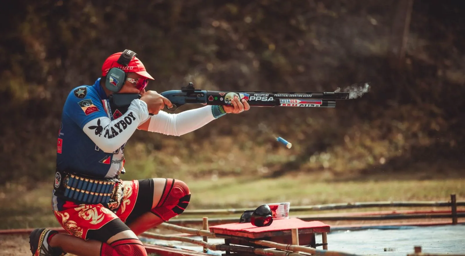 Diz çökmüş sporcu pompalı yivsiz tüfekle atış yapıyor; boş kovan havada
